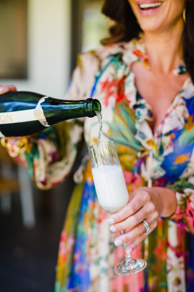 Woman in a bright patterned maxi dress pouring a bottle of champagne into a glass while laughing