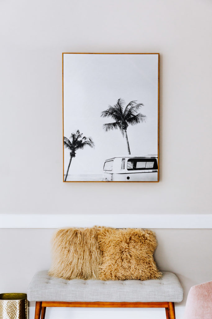 Canvas with palm tree design hanging on wall above bench holding two furry yellow pillows
