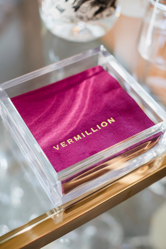 Close up image of a stack of fuscia pink napkins with the word Vermillion written in gold letters sitting inside an acrylic napkin holder on a glass top table | Sara Coffin Photography