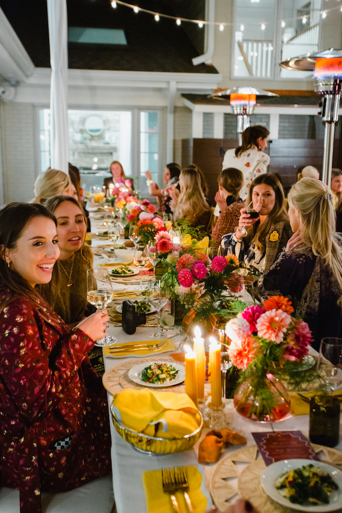 Eleven women sit at long banquet table decorated with candles and florals outdoors under space heaters while conversing with drinks in hand | Sara Coffin Photography for a Small Business Photo Shoot