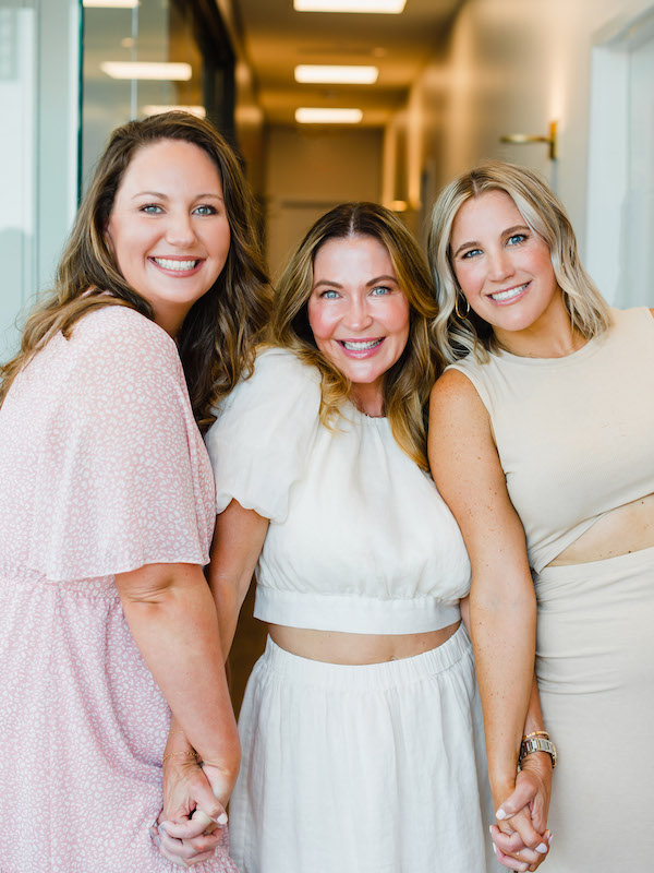 Three women hold hands and smile at the camera while standing in hallway during a small business photo shoot | Sara Coffin Photography