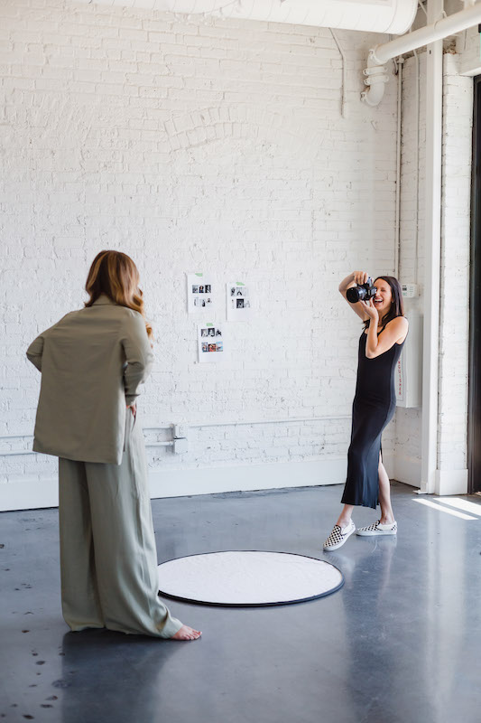 Photographer in a black dress taking photos of a woman wearing a casual sage green two piece suit inside a large empty photography studio | Sara Coffin Photography