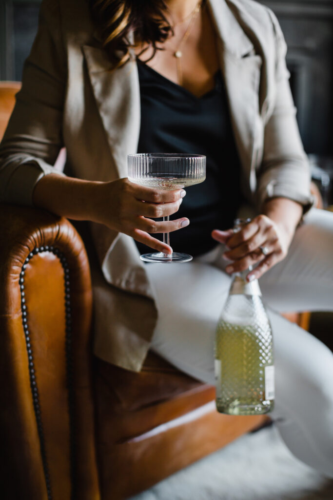 Woman holds martini glass and bottle of champagne while sitting on a camel colored leather chair | Personal branding photography session for Abigail Jackson Photography by Sara Coffin Photo