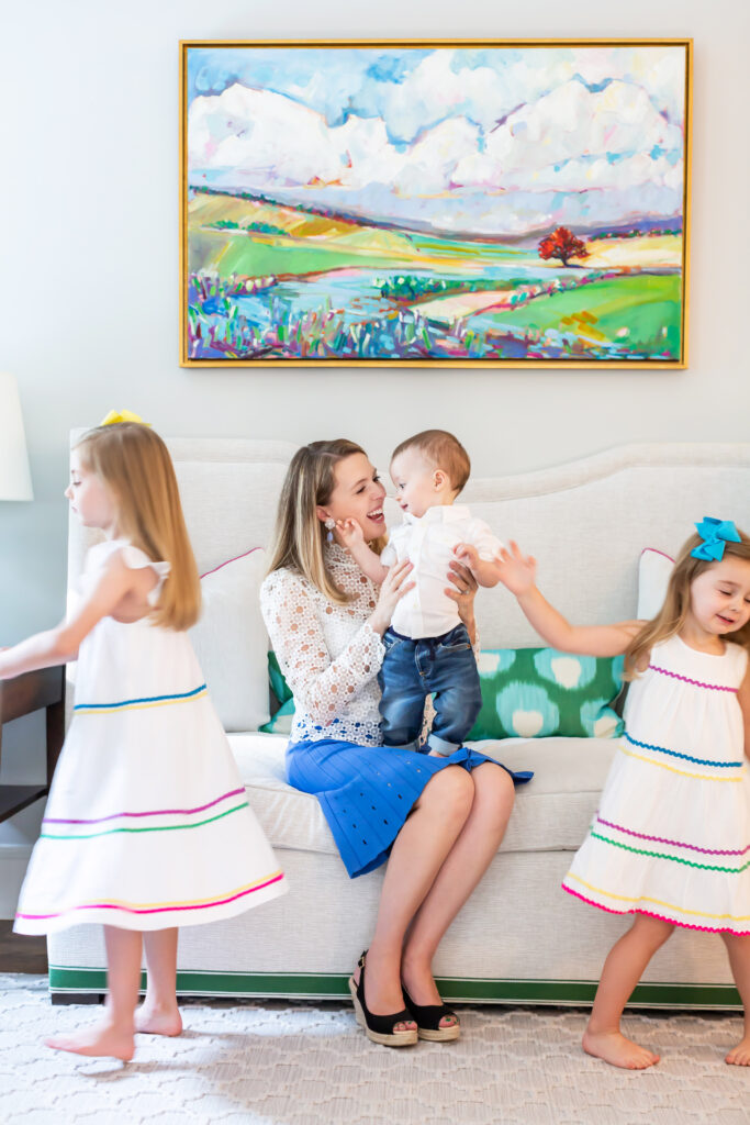 Woman in a blue skirt sits on a couch holding a baby on her lap while two girls in striped dresses run through the room | Personal branding session for Interior Designer Lauren McKay Interiors by Sara Coffin Photo