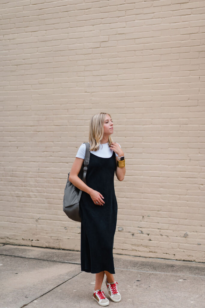 Girl in black slip dress poses with backpack slung on one shoulder in front of a blank brick wall in downtown Southern Pines NC