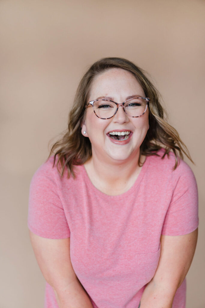Woman in a pink shirt and glasses laughs while looking into the camera