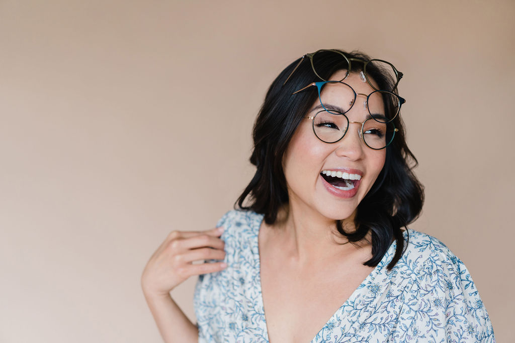 Dr. Amy Kasper of The Vision Studio in Cary, NC wearing three pairs of glasses stacked on her face while laughing in front of a tan backdrop | Sara Coffin Photo