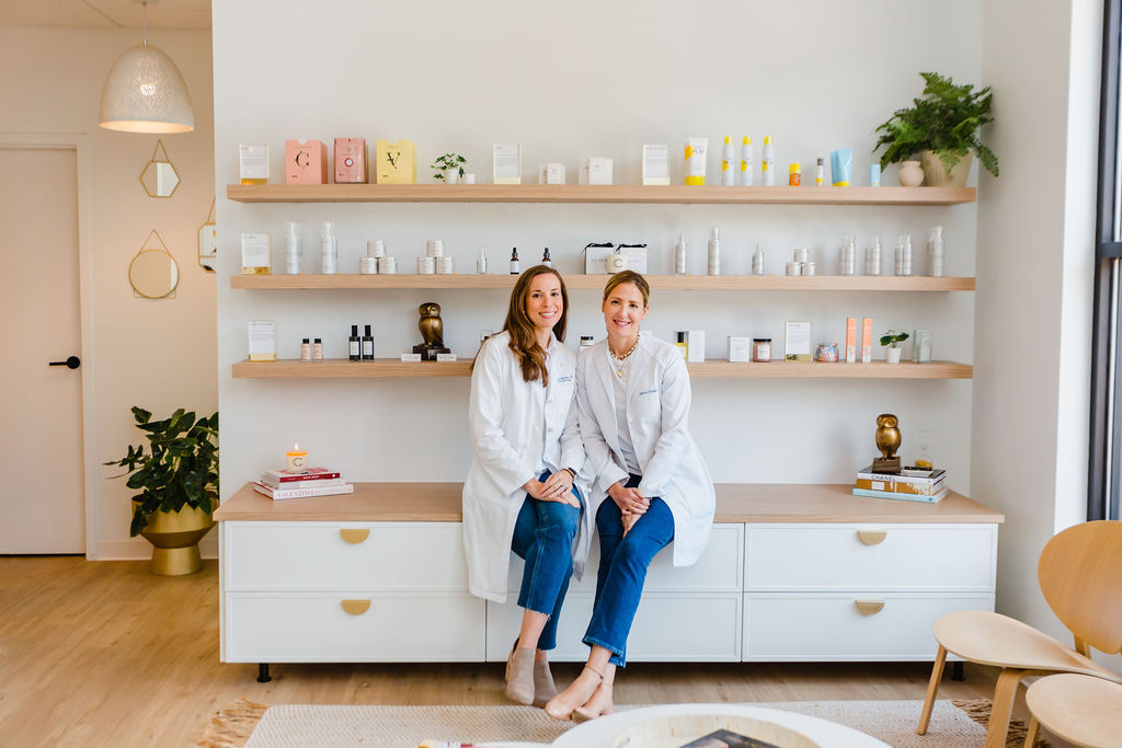 Doctor Bialas and Doctor Hooten of Clara Dermatology sit next to each other in front of skincare retail shelf during personal branding photography session