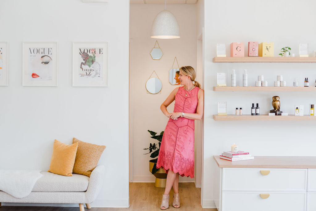 Woman in a pink eyelet lace midi dress leans against a wall holding floating shelves of skincare products at Clara Dermatology office in Cary NC