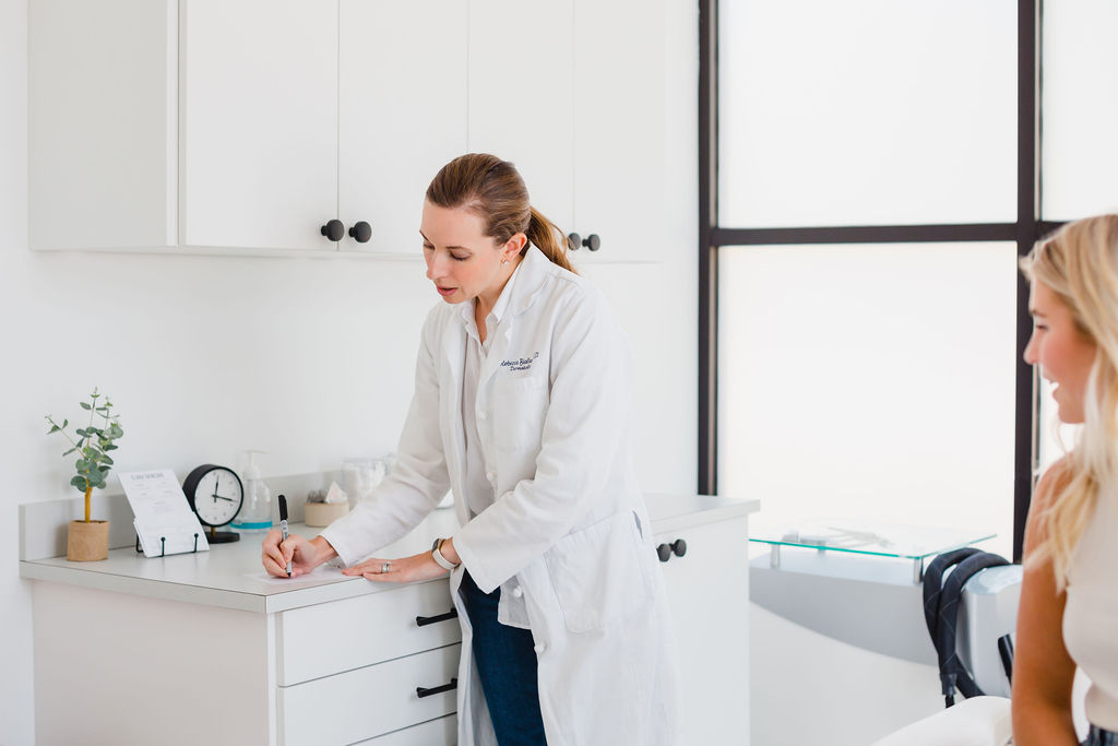 Female dermatologist in a white lab coat leans over a countertop to look at a piece of paper