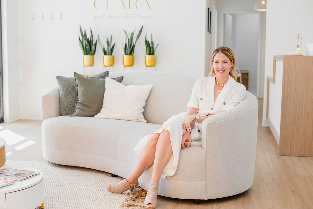 Dr. Hooten of Clara Dermatology in Cary, NC wears a white dress and leans back on a linen couch in waiting area 