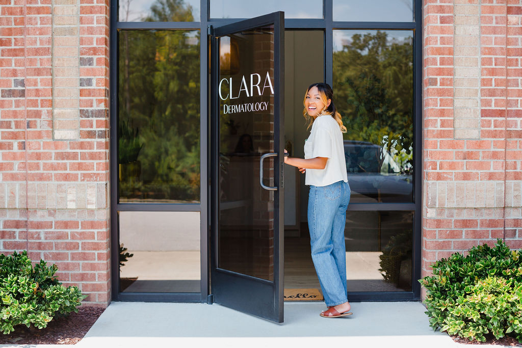 Woman in a white t-shirt and blue jeans smiles as she walks through the front door to Clara Dermatology in Cary, NC