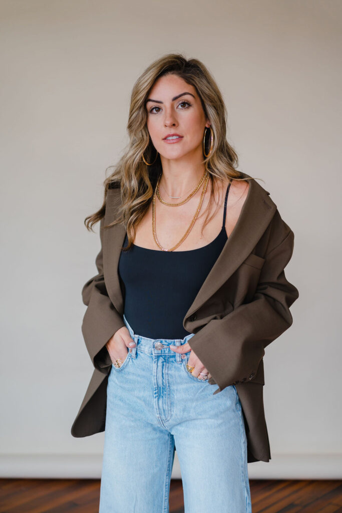 Model wears light wash jeans and black bodysuit with an oversized olive green blazer