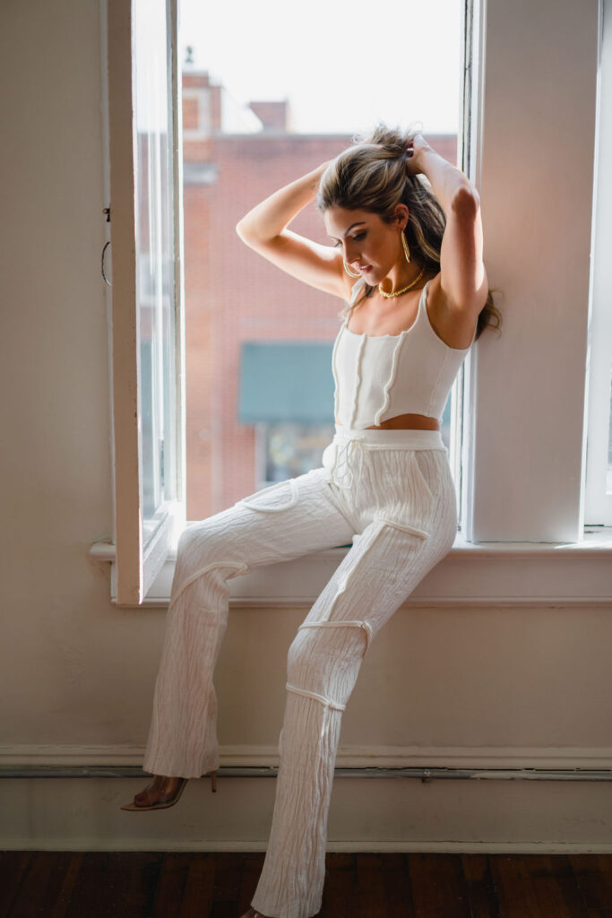 Model wears white drawstring patchwork pants with a white cotton corset top while sitting on the window sill of an open window in an empty photo studio