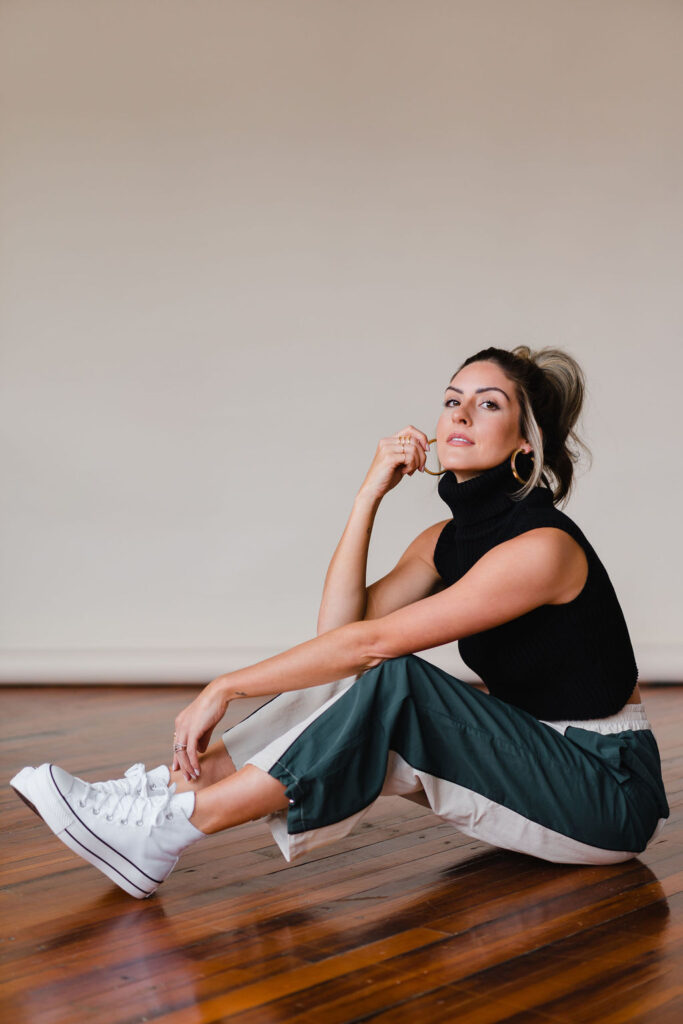 Model wears green and white sweatpants and black cowl neck sweater vest with white high top converse while sitting on the floor in front of a tan photo backdrop in a photo studio