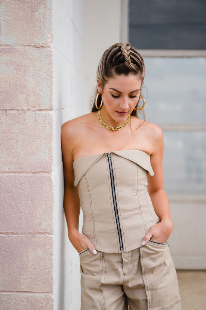 Model wears a beige matching set with a zip front tube top and wide legged pants and gold hoop earrings with a braided half up hairstyle