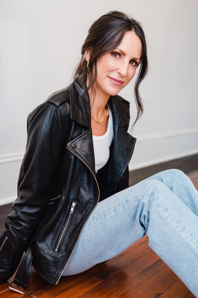 Woman wears light wash jeans and black leather jacket while sitting on the floor and smiling at the camera