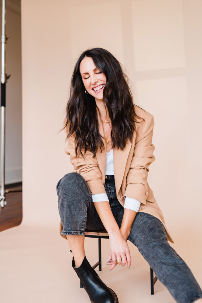 Woman with dark brunette hair wears acid wash black jeans and a camel colored blazer and laughs while sitting on a low stool in front of a tan photo backdrop