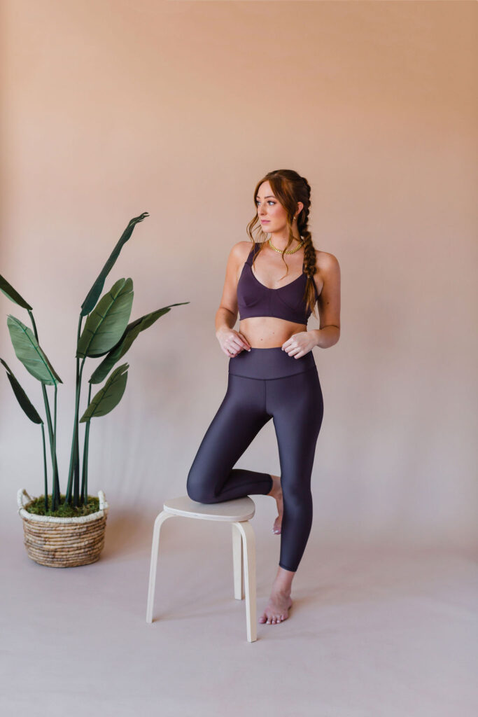 Woman wears a purple matching workout set from Alo Yoga while resting one knee on a light wood stool