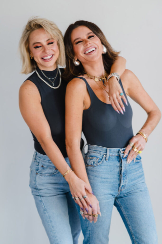Two women wearing black bodysuits and light wash jeans lean against each other and laugh while holding hands