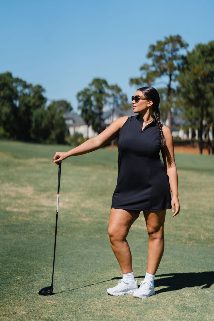 Woman in black golf dress and sunglasses leans on her putter on a golf course | Branding photography for Harlow Sport