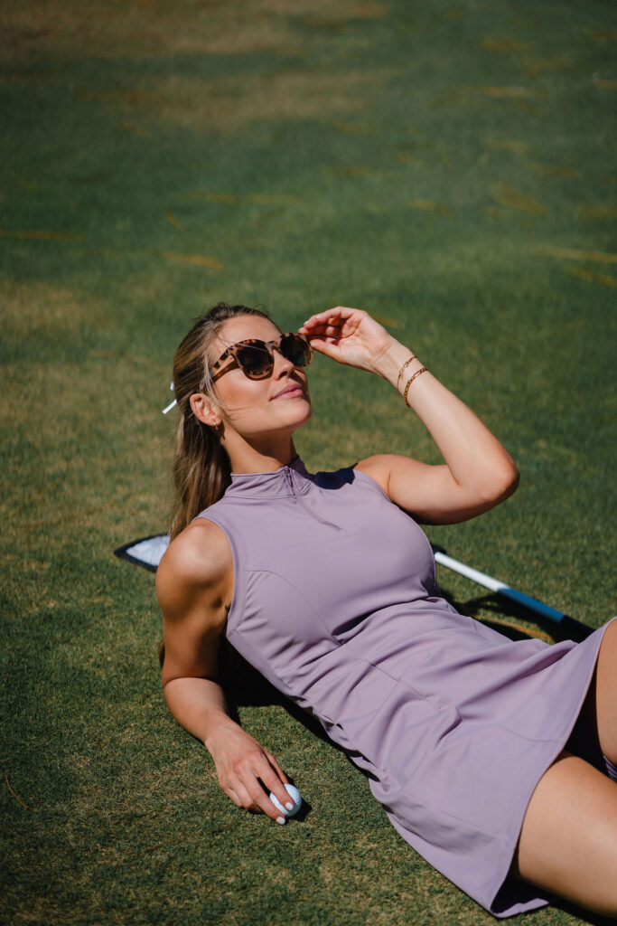 Woman in purple golf dress and tortoise sunglasses lays on golf course