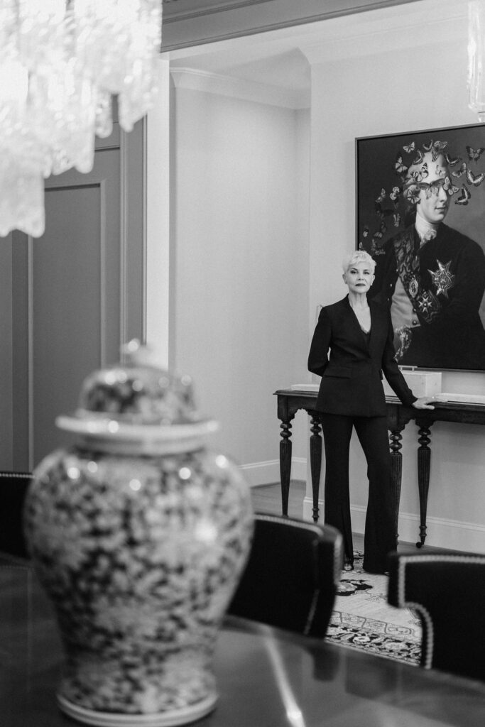 Woman in a black two piece suit rests her hand on a hallway table underneath a large hanging portrait | Personal branding photoshoot for realtor Marianne Mansour