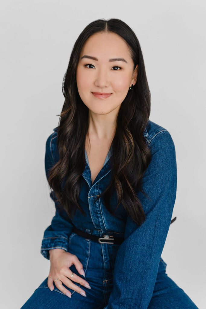 Asian woman wears a dark wash denim jumpsuit and sits in front of a white backdrop in a photography studio during a professional headshot session | Sara Coffin Photo Headshot Mini Marathon session experience