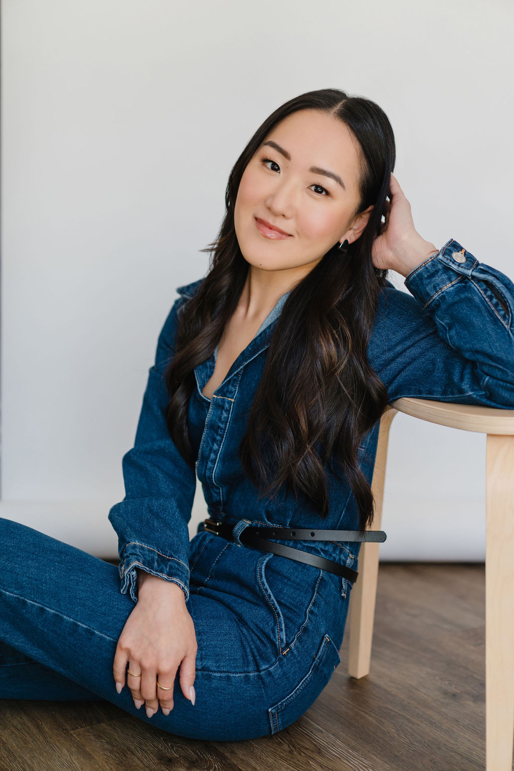 Asian woman wearing a dark wash denim jumpsuit leans back against a chair in front of a white backdrop in a photography studio during a professional portrait session