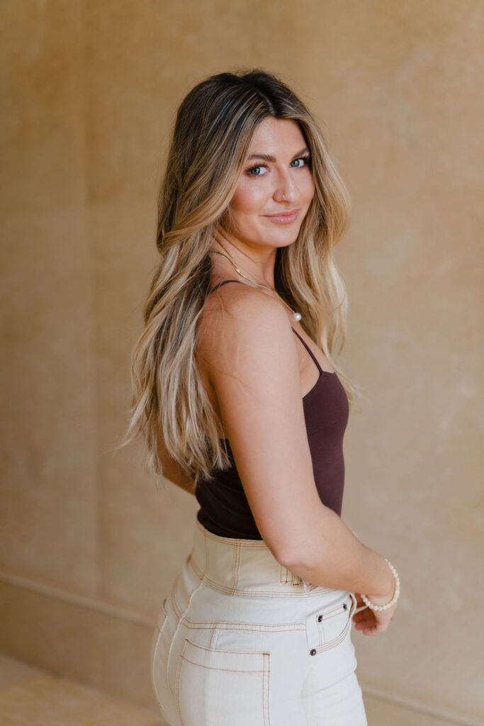 Blonde woman in brown bodysuit and tan cargo pants outfit smiles at the camera in front of a tan marble backdrop at Oxbow Estate in Clayton, NC during Sara Coffin Photo branding photoshoot