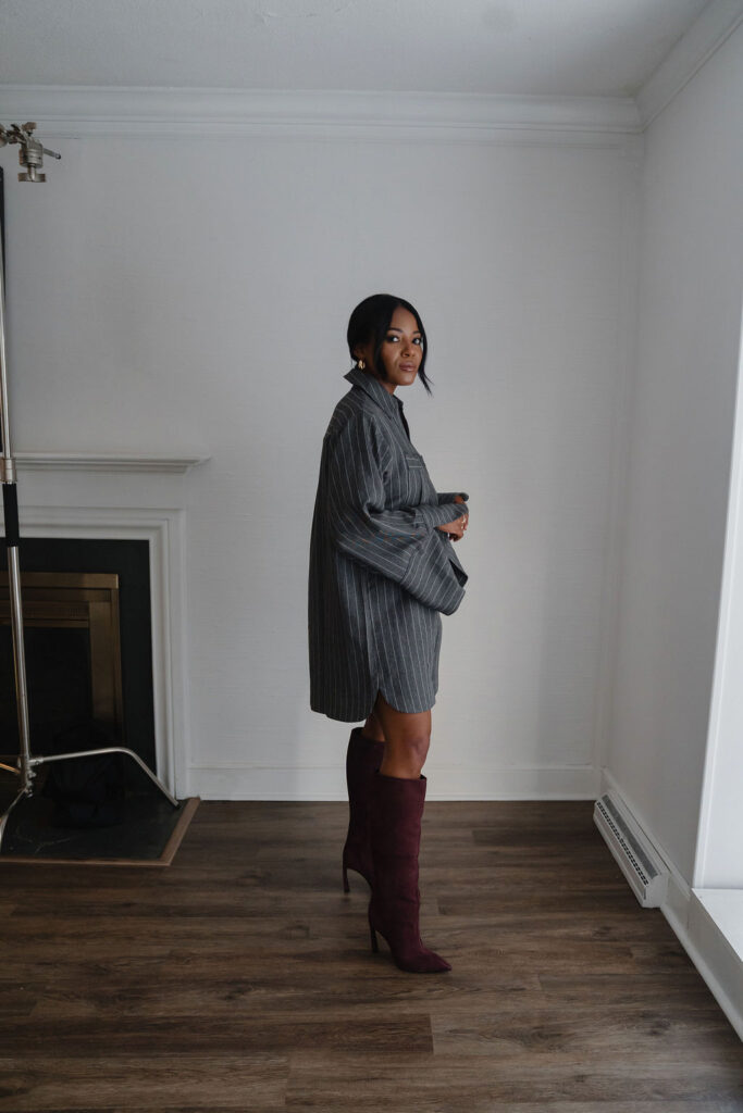 Black female entrepreneur wears charcoal gray pinstripe oversized shirt dress with maroon suede boots and poses inside a North Carolina photo studio | Sara Coffin Photo