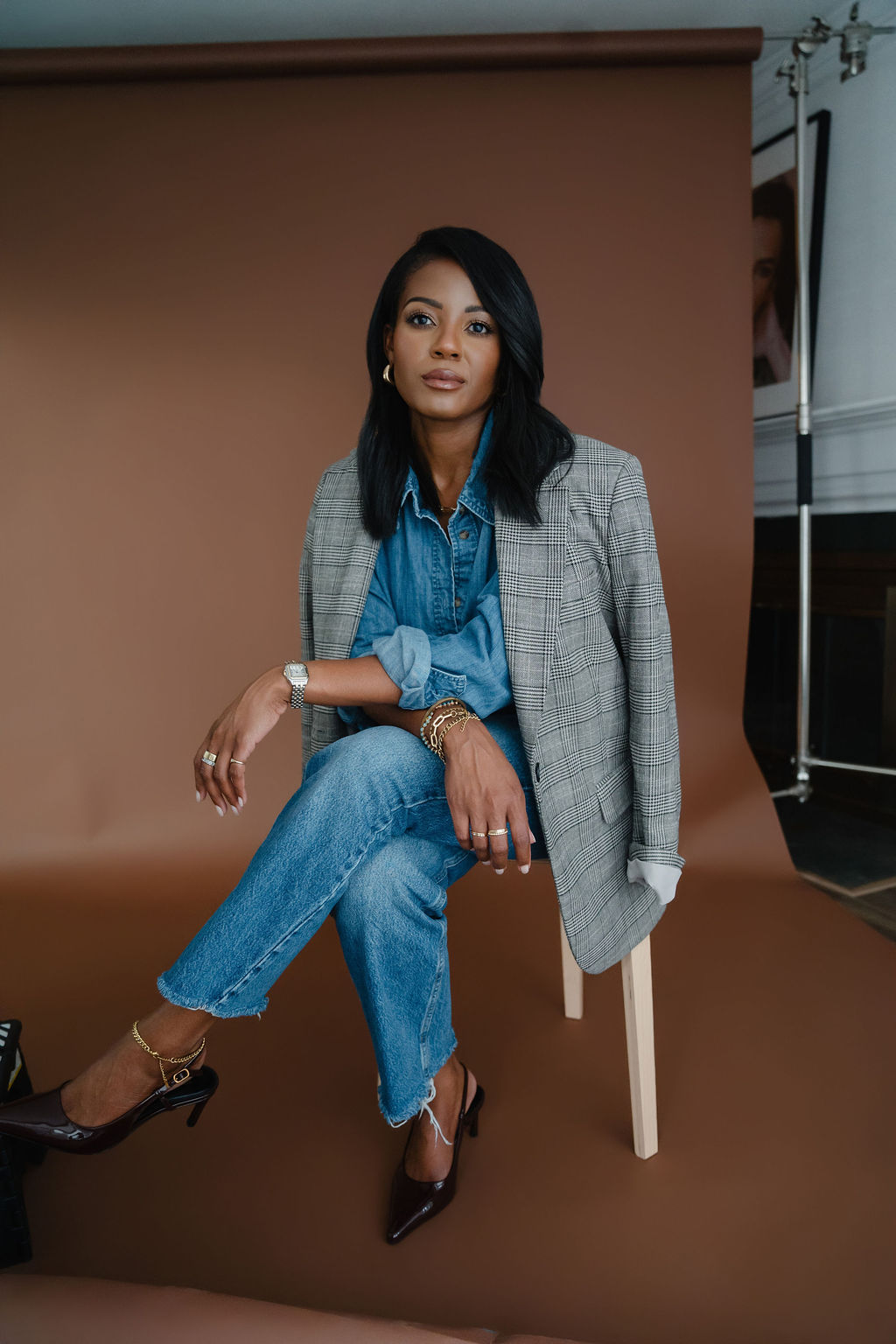 Black female entrepreneur sits on a wooden stool in a gray plaid blazer and denim shirt combo with straight leg jeans in front of a brown backdrop inside a North Carolina photo studio | Sara Coffin Photo