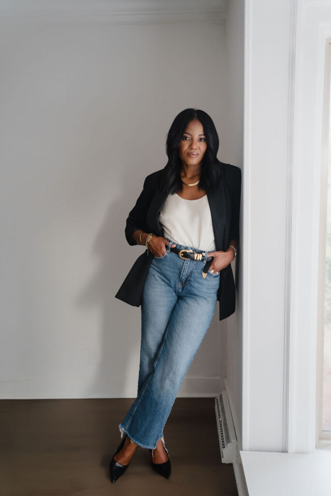 Black female entrepreneur wears a black blazer and white shirt combo with straight leg jeans and leans against the wall inside a North Carolina photo studio | Sara Coffin Photo