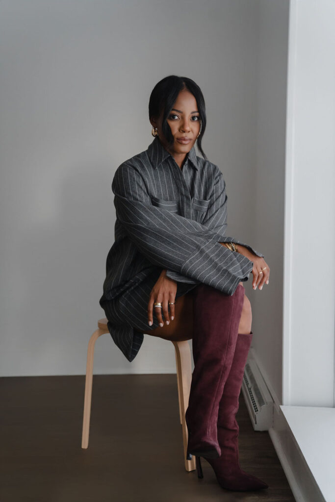 Black female entrepreneur wearing charcoal gray pinstripe oversized shirt dress and maroon suede boots sits on a wooden stool and poses for portraits inside a North Carolina photo studio | Sara Coffin Photo