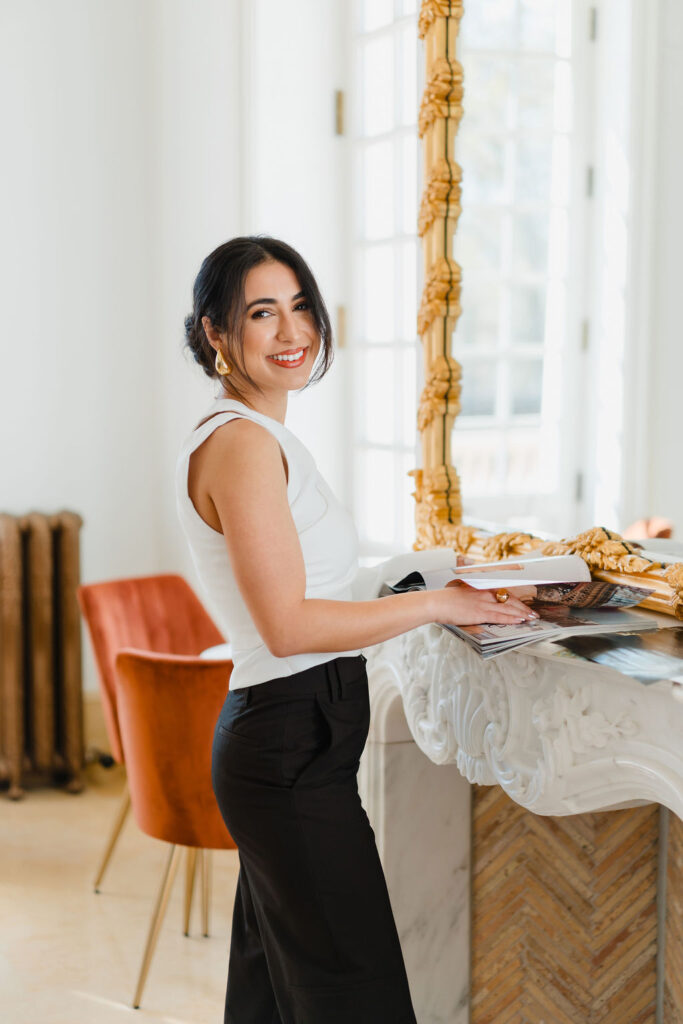 Brunette woman in white shirt and brown trousers poses at an intricate mantelpiece flipping through a magazine and smiling at the camera during a branding photoshoot by Sara Coffin Photo at Oxbow Estate in Clayton, NC | Photoshoot hair and makeup tips